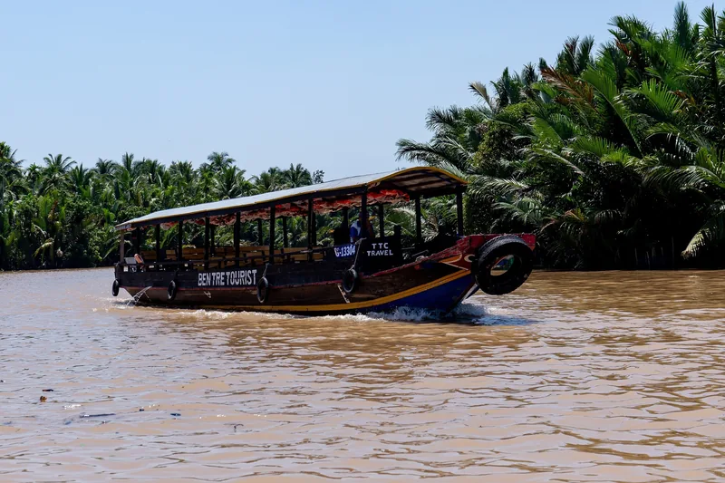 A wooden tourist boat cruises along a muddy river lined with coconut palms in the Mekong Delta near Ben Tre.