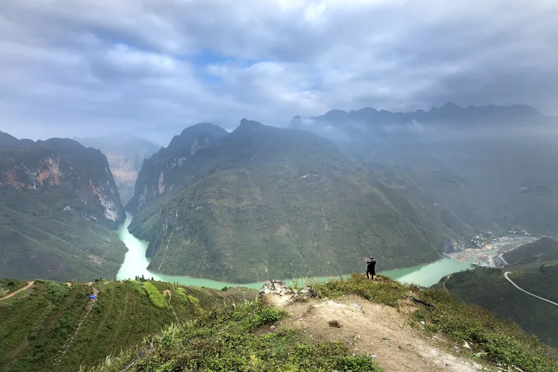 A person stands on a cliff overlooking the turquoise Nho Que River winding through steep limestone mountains in Ha Giang. A person stands on a cliff overlooking the turquoise Nho Que River winding through steep limestone mountains in Ha Giang.