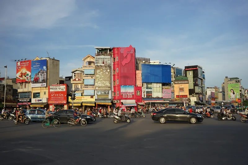 Busy Hanoi intersection with cars and motorbikes passing colorful shopfronts and billboards.