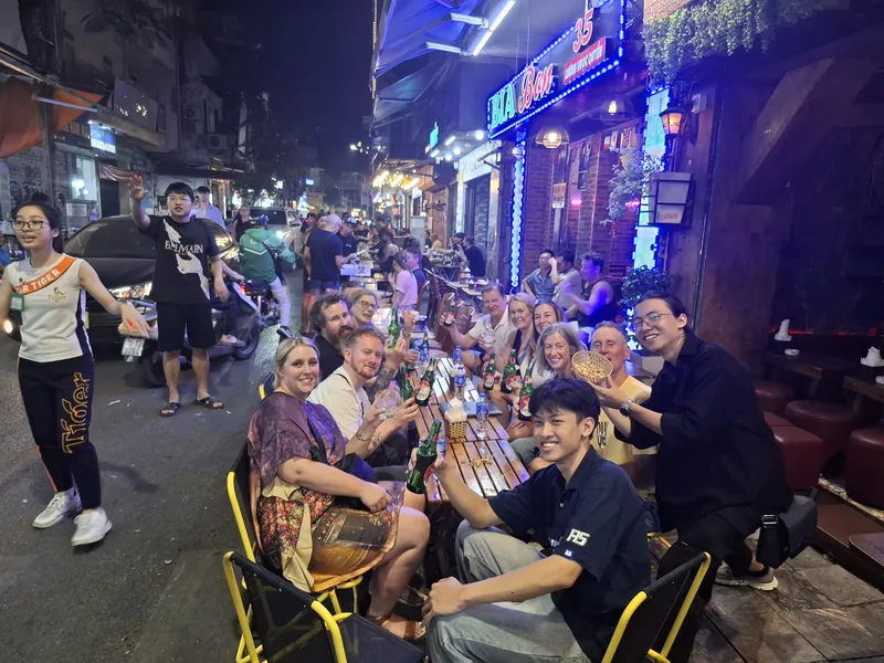 Tourists share beers at low plastic tables in Hanoi's bustling beer street at night.