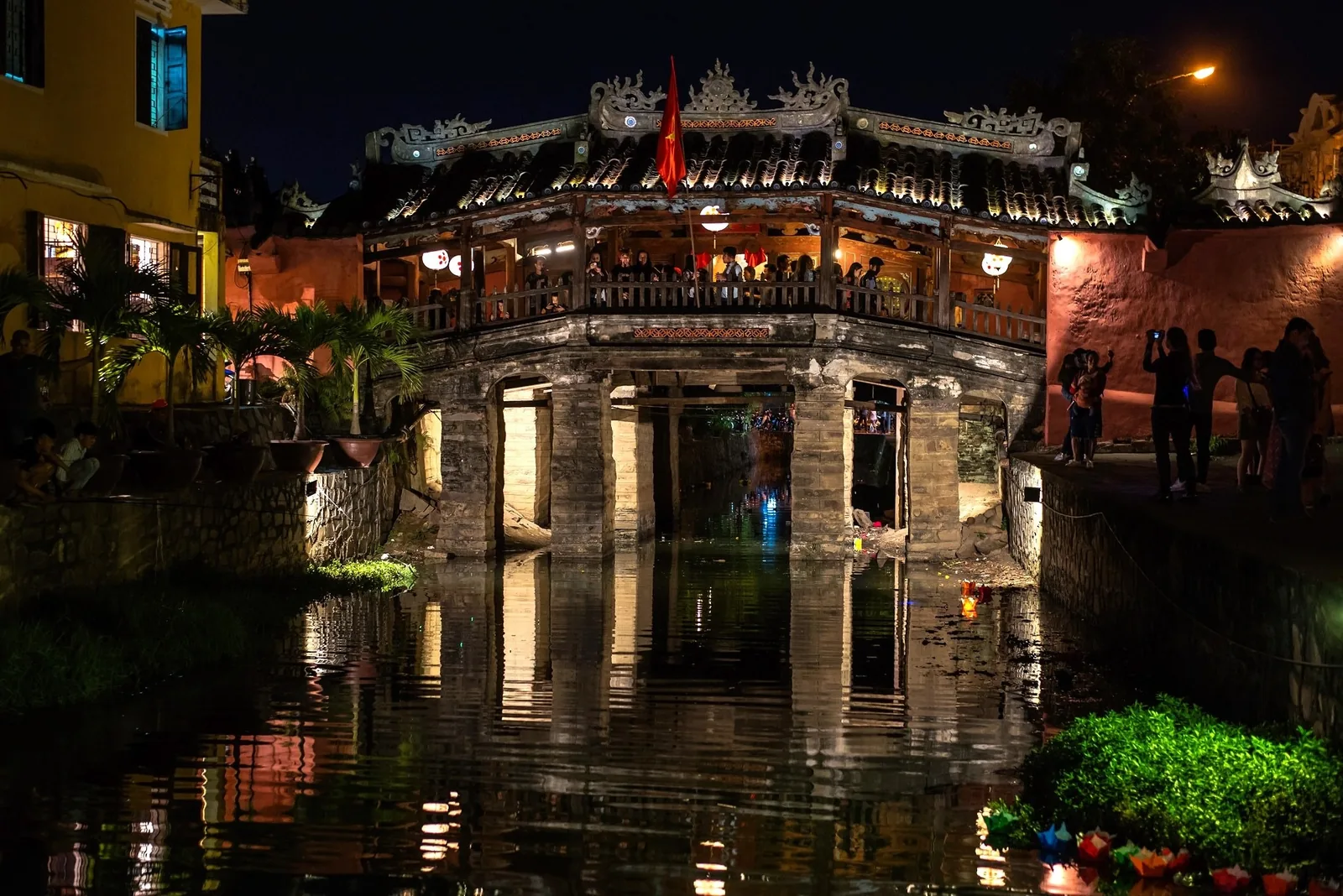 The Japanese Covered Bridge glows at night with tourists crossing above its reflection in Hoi An's canal. The Japanese Covered Bridge glows at night with tourists crossing above its reflection in Hoi An's canal.