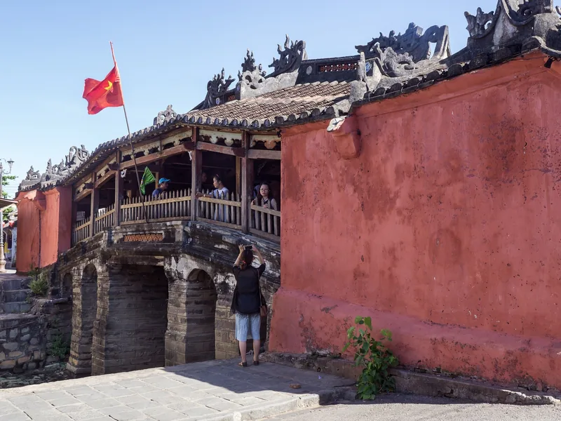 Tourists cross the wooden Japanese Covered Bridge beside a weathered ochre wall in Hoi An's old town. Tourists cross the wooden Japanese Covered Bridge beside a weathered ochre wall in Hoi An's old town.