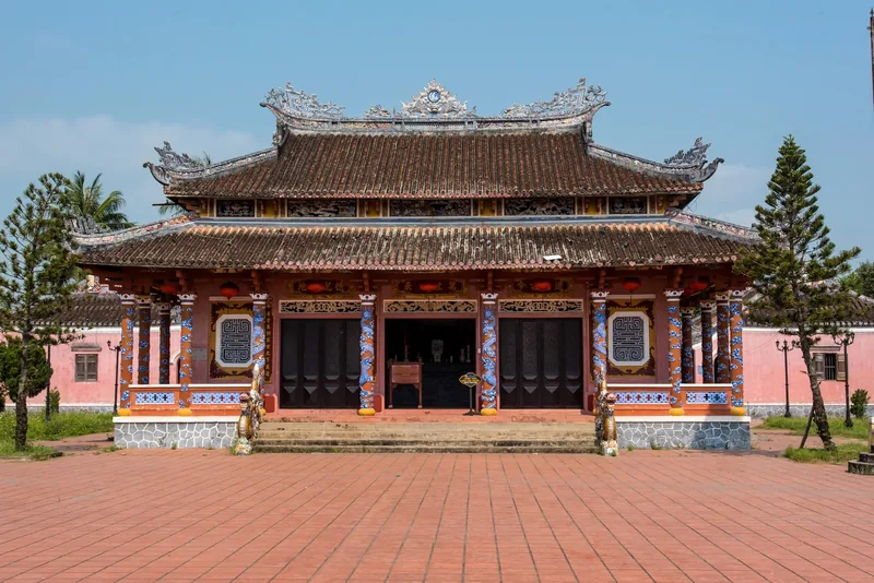 Ornate tiled roof and mosaic columns decorate a traditional communal house in Hoi An under a clear sky. Ornate tiled roof and mosaic columns decorate a traditional communal house in Hoi An under a clear sky.