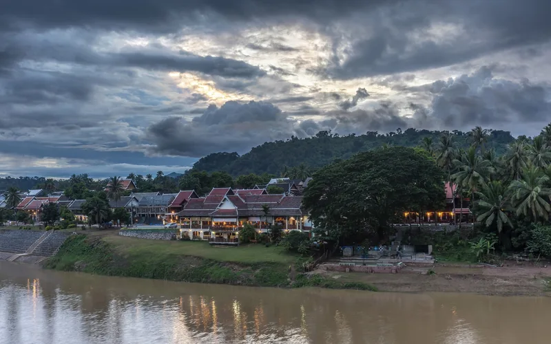 Illuminated riverside buildings and lush hillside under a dramatic dusk sky along the Nam Khan River in Luang Prabang.
