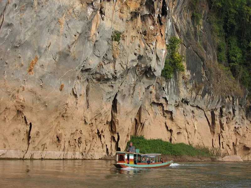 A wooden longtail boat cruises past towering limestone cliffs on the Nam Ou River near Nong Khiaw.
