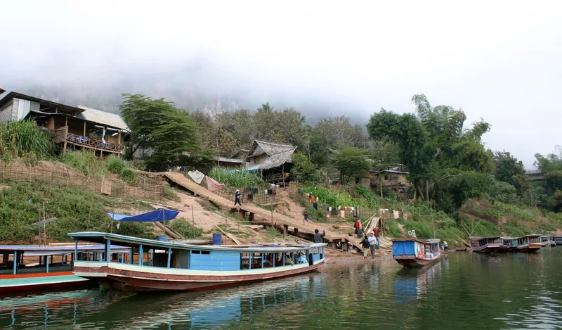 Wooden boats moored along the riverbank at Nong Khiaw, with village houses and misty hills behind.