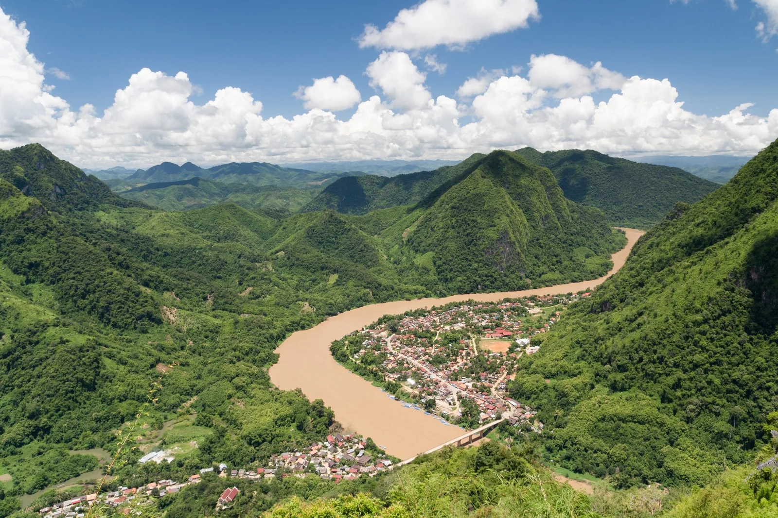 A muddy river bends through a small riverside town surrounded by lush green mountains near Nong Khiaw, Laos.