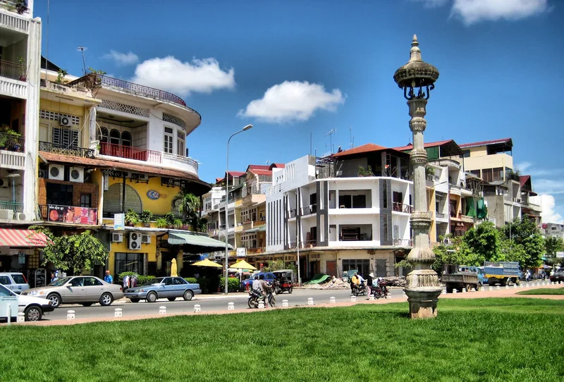 A row of colorful French-colonial style buildings lines a busy street in Phnom Penh, with a lamp post and green lawn in the.