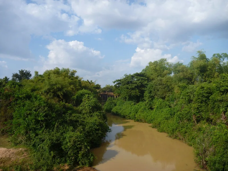 A muddy river winds past lush greenery near an old bridge in the Siem Reap countryside.