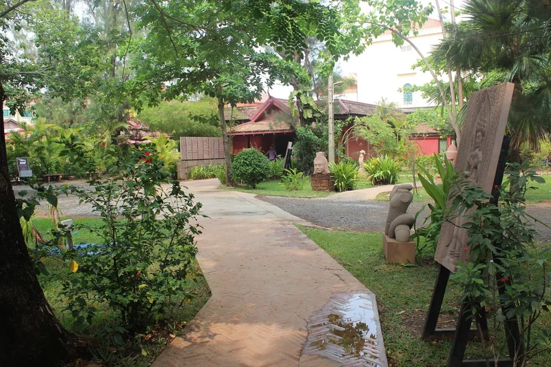 A shaded garden path leads past stone carvings and traditional wooden buildings at Artisans Angkor in Siem Reap.