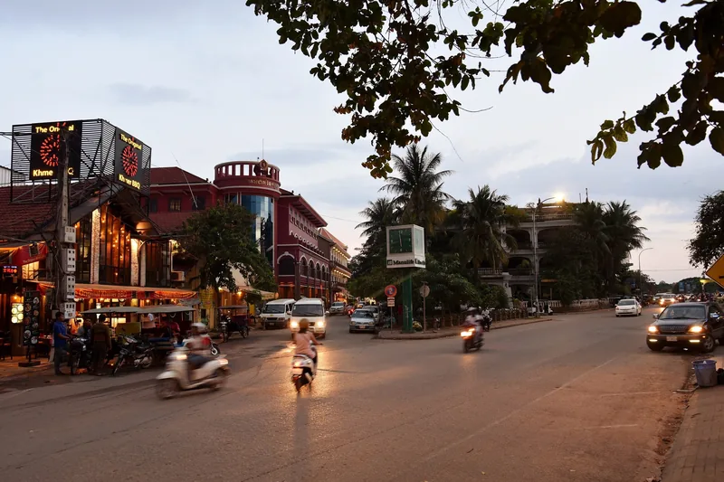 Motorbikes and cars travel a busy street in Siem Reap at dusk, with restaurants and palm trees lining the road.
