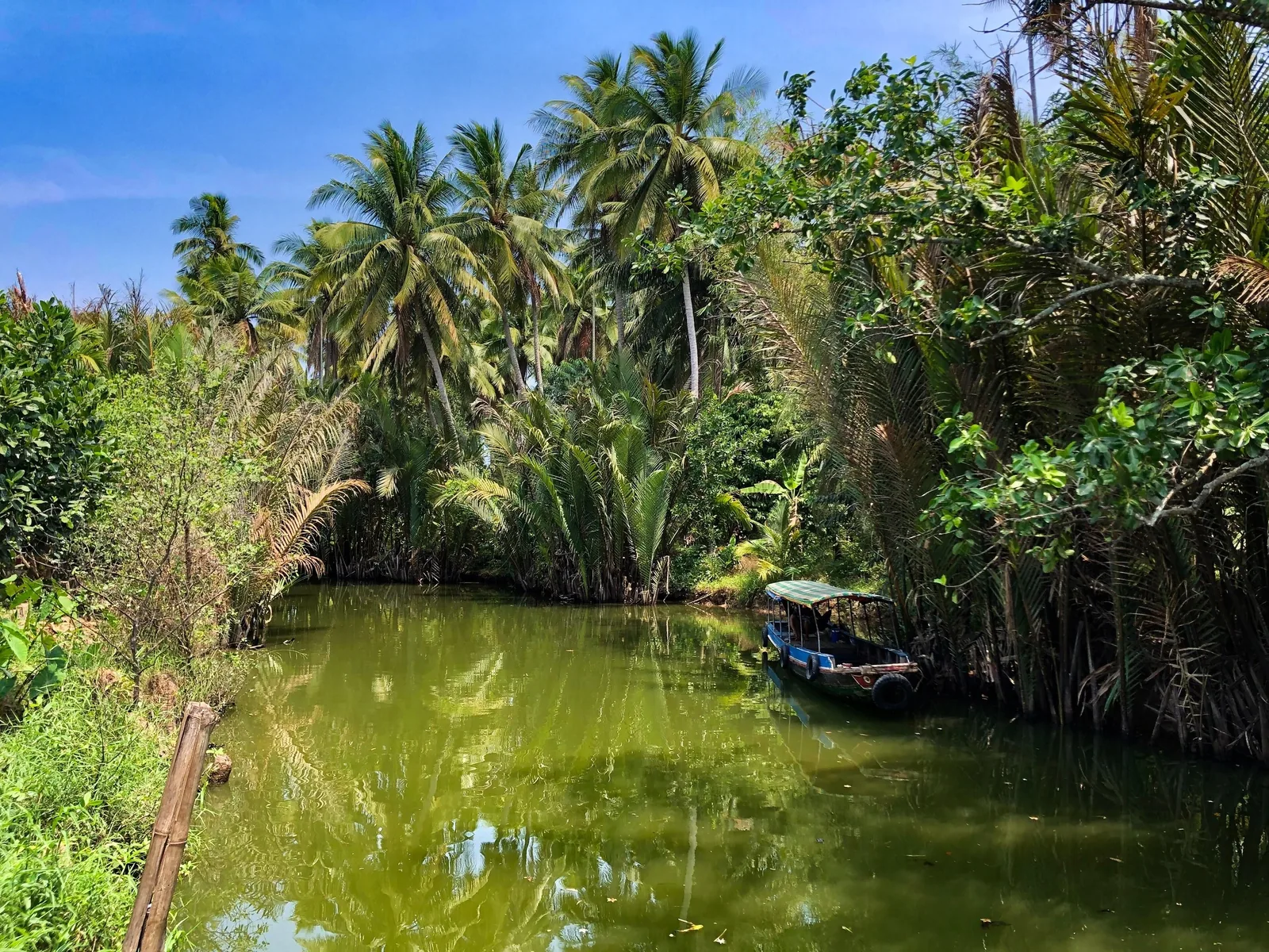 A wooden canopy boat rests on a calm green canal lined with coconut palms in the Mekong Delta.
