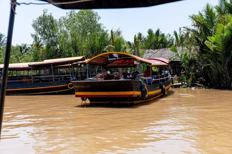 A wooden tourist boat with a red canopy carries passengers along the muddy waters of the Mekong Delta.