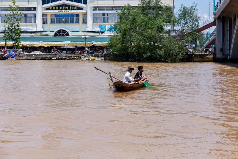 Two people paddle a small wooden boat on the muddy river in front of Ben Tre market, Mekong Delta.