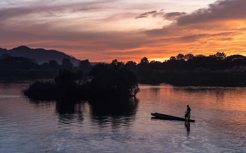 A fisherman stands on his boat silhouetted against a fiery sunset sky over the Mekong Delta river.