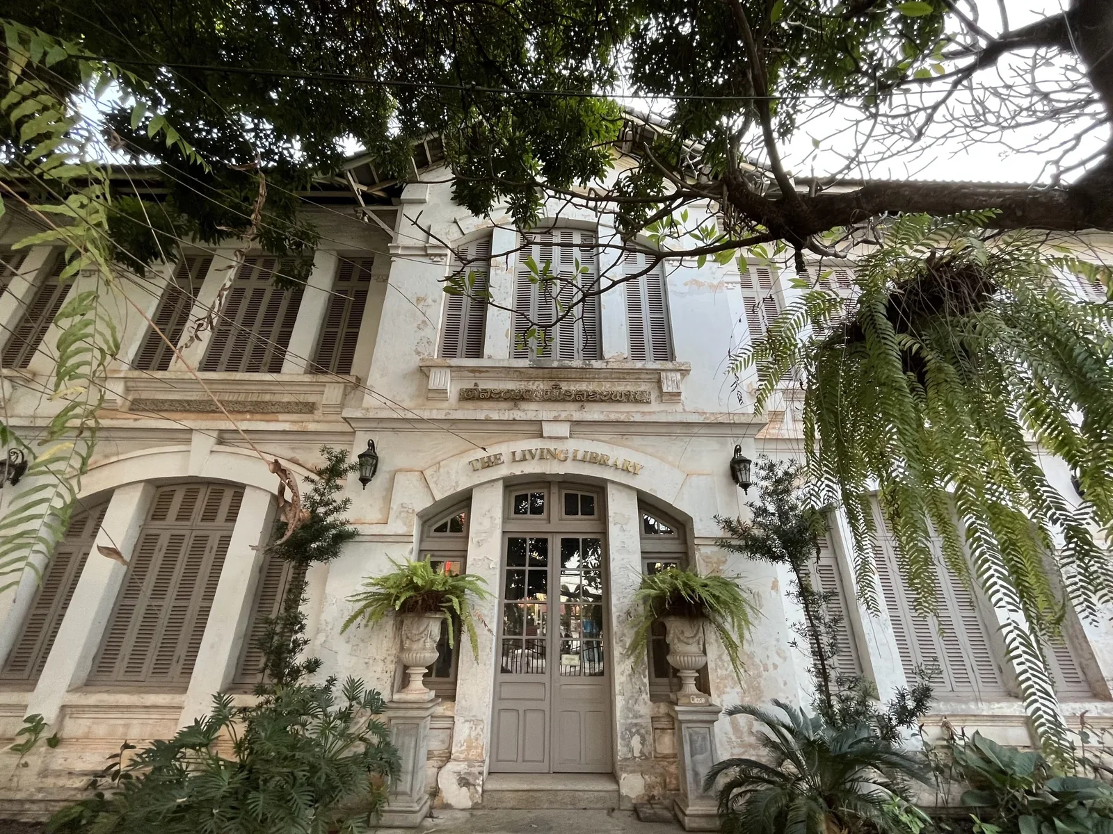 A weathered French colonial building with shuttered windows and hanging ferns in Vientiane, Laos.